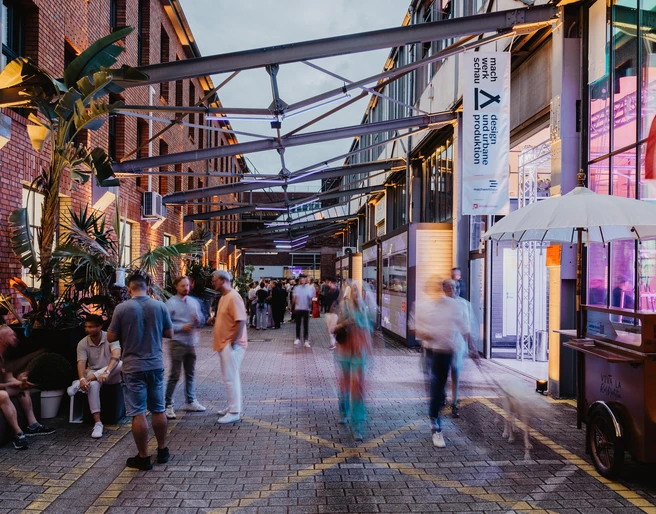 Menschen flanieren auf einem beleuchteten Innenhof mit roten Backsteingebäuden und tropischen Pflanzen.People stroll around an illuminated inner courtyard with red brick buildings and tropical plants.
