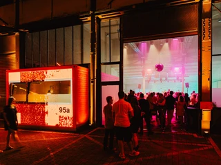 Anhand der Bildanalyse kann folgende Beschreibung für das alt-Attribut erstellt werden: Besucher auf einem beleuchteten Innenhof, im Hintergrund eine Partylocation mit bunter Lichtshow.Based on the image analysis, the following description can be created for the alt attribute: Visitors in an illuminated courtyard, in the background a party location with a colorful light show.