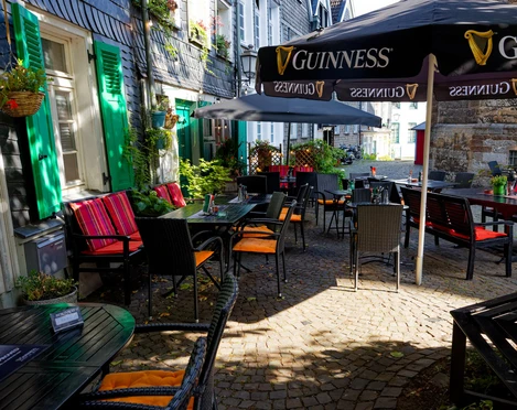 Biergarten, Dingle`s Irish Pub in Mettmann Ein Biergarten mit dunklen Holztischen und roten Sitzauflagen unter Guinness-Sonnenschirmen.