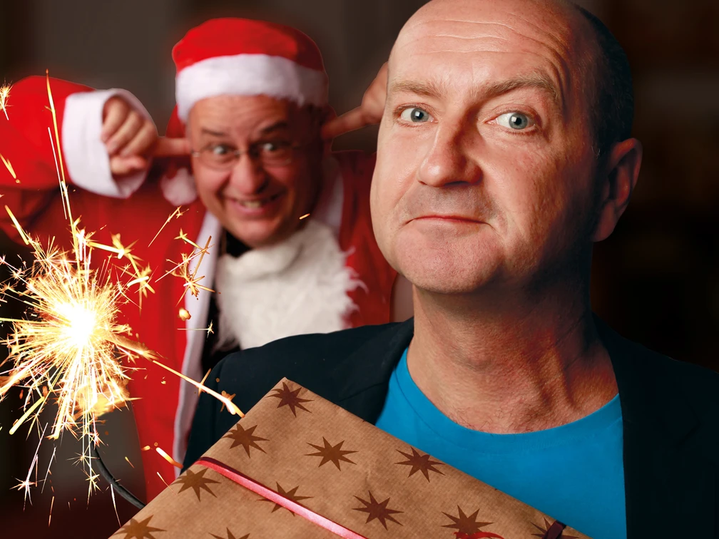 251206_SchwGruetze.jpg Ein Mann hält ein Geschenk vor seinem Oberteil, während im Hintergrund eine Person im Weihnachtsmannkostüm mit einem schalkhaften Ausdruck die Ohren zuhält. Eine Wunderkerze sprüht funkelndes Licht.