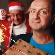 251206_SchwGruetze.jpg Ein Mann hält ein Geschenk vor seinem Oberteil, während im Hintergrund eine Person im Weihnachtsmannkostüm mit einem schalkhaften Ausdruck die Ohren zuhält. Eine Wunderkerze sprüht funkelndes Licht.