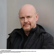 Tad Williams Ein breitschultriger Mann mit Glatze und Bart, in dunkler Kleidung, lehnt an einer Metallwand.A broad-shouldered man with a bald head and beard, wearing dark clothes, is leaning against a metal wall.