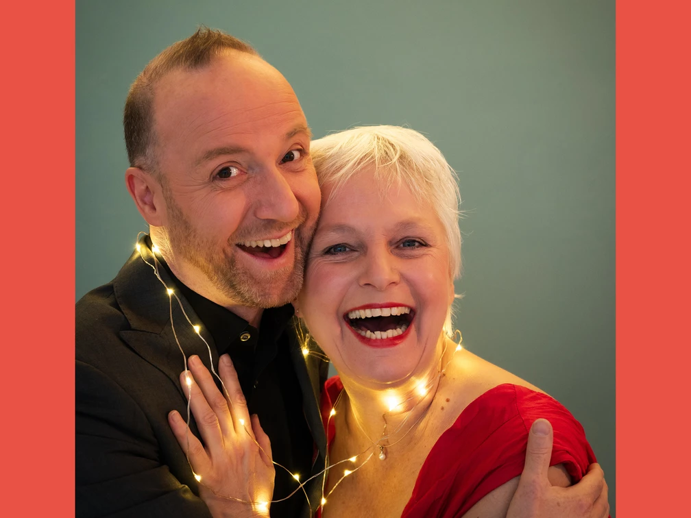 251216_Lametta.jpg Ein fröhliches Paar mittleren Alters lächelt, umgeben von einer Lichterkette vor grünem Hintergrund.