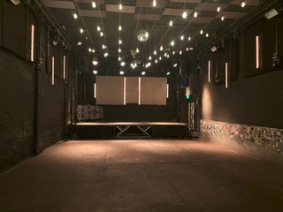 Helio37 Mainfloor Leerer Veranstaltungsraum mit Bühne, umgeben von dunklen Wänden und Lichterketten an der Decke.Empty event room with stage, surrounded by dark walls and fairy lights on the ceiling.