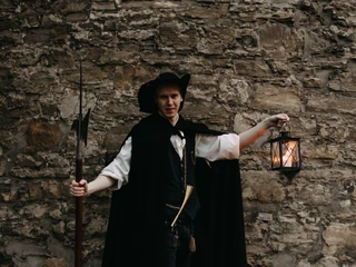 Beschwipster Nachtwächter (2).jpg Ein Nachtwächter in traditioneller Kleidung lehnt an einer alten Steinmauer und hält eine Laterne.