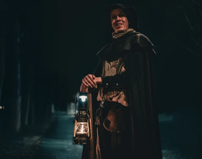 Galgen, Gräber & Ganoven (Hauptbild).jpg Ein Nachtwächter in traditionellem Gewand hält eine Laterne und steht auf einem dunklen Weg.