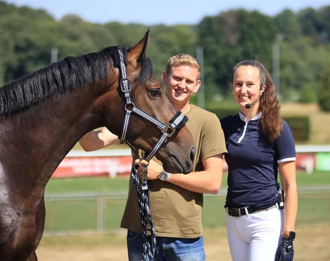 Zwei Menschen stehen lächelnd neben einem Pferd auf einem Rasenplatz an einem sonnigen Tag.