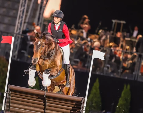 Reiterin springt mit einem braunen Pferd über ein Hindernis, im Hintergrund ein Orchester.