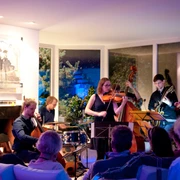 Musik in den Häusern der Stadt 2017 Eine Gruppe von Musiker*innen spielt konzentriert in einem modern beleuchteten Raum vor einem sitzenden Publikum.A group of musicians plays in a modern, well-lit room in front of a seated audience.