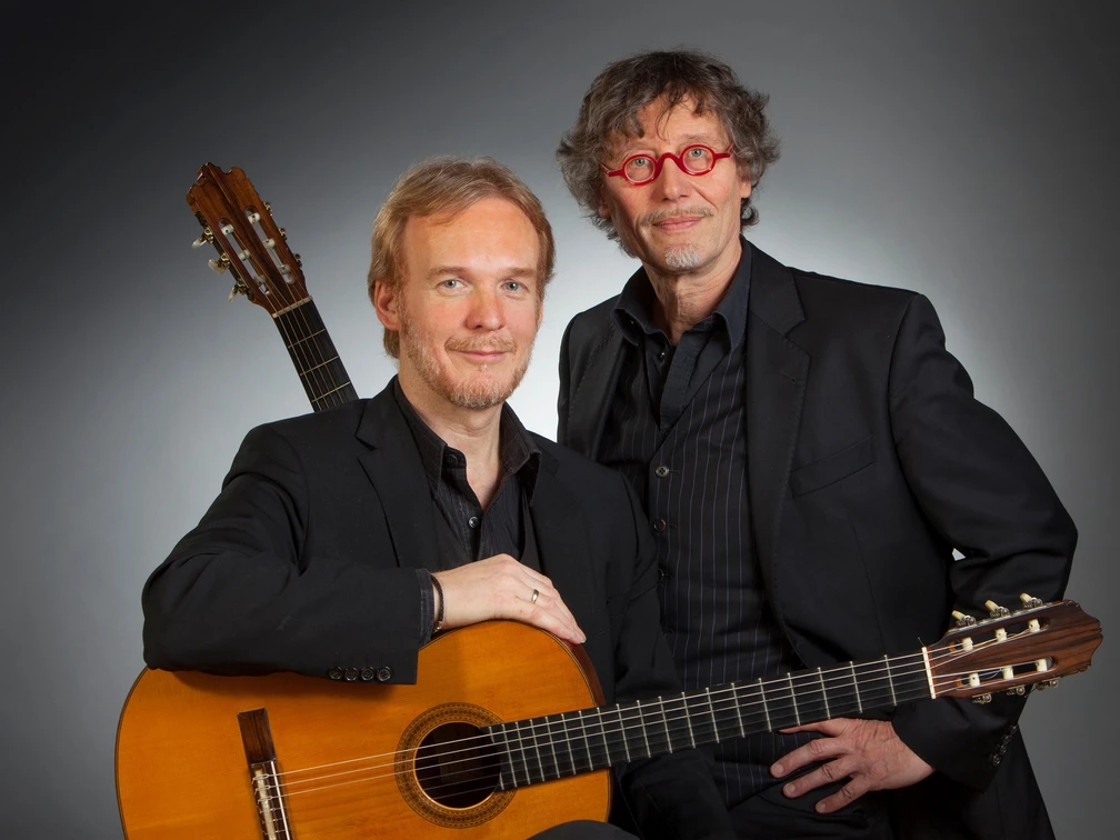Bernd Steinmann & Stefan Loos Zwei Musiker mit klassischen Gitarren posieren in dunkler Kleidung vor neutralem Hintergrund.Two musicians with classical guitars pose in dark clothing in front of a neutral background.