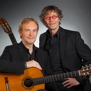 Bernd Steinmann & Stefan Loos Zwei Musiker mit klassischen Gitarren posieren in dunkler Kleidung vor neutralem Hintergrund.Two musicians with classical guitars pose in dark clothing in front of a neutral background.