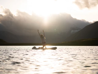 Sonnenuntergangs-Yoga auf dem See Frau macht SUP Yoga Bettmersee im Sommer bei Sonnenuntergang mit goldener Spiegelung auf der Wasseroberfläche und Bergen im Hintergrund