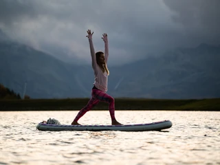 Yoga auf SUP bei Abendstimmung SUP Yoga Bettmersee im Sommer mit Frau in Kriegerhaltung bei stimmungsvoller Abendsonne und Bergsilhouette im Hintergrund