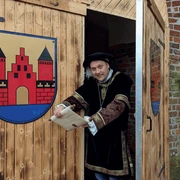 Markteröffnung.png Mann in historischer Kleidung öffnet Holztor mit Stadtwappen und hält eine Schriftrolle in der Hand.
