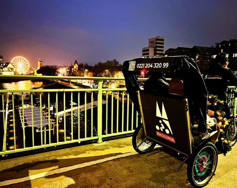 RikoloniaWinterTeamlicht.png Ein winterlich beleuchtetes Rikscha-Team vor dem Kölner Rheinpanorama bei Abenddämmerung.A wintery illuminated rickshaw team in front of the Cologne Rhine panorama at dusk.