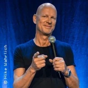 Portrait des lächelnden Mannes mit Mikrofon auf einer Bühne, vor blauem Hintergrund.