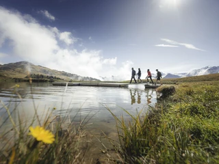 Foxtrail hike along the lakeshore Foxtrail Desoria Aletsch Arena mit vier Personen, die über einen Steg am Bettmersee wandern, vor den Walliser AlpenFoxtrail Desoria Aletsch Arena with four people hiking over a footbridge on Lake Bettmersee, in front of the Valais AlpsFoxtrail Desoria Aletsch Arena avec quatre personnes marchant sur une passerelle au Bettmersee, devant les Alpes valaisannes