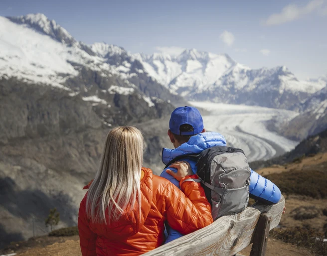 Aussicht von der Hohfluh Frühlingsfahrten Hohfluh mit einem Paar in bunten Jacken auf einer Bank mit Blick auf den Grossen AletschgletscherSpring rides Hohfluh with a couple in colorful jackets on a bench with a view of the Great Aletsch GlacierCourses printanières à Hohfluh avec un couple en vestes colorées sur un banc avec vue sur le grand glacier d'Aletsch
