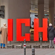 ICH.!!! - Installation in public space Rote Skulptur "ICH" vor moderner Architektur, betrachtet von zwei Passanten. Urbanes Straßenbild.Red sculpture "ICH" in front of modern architecture, viewed by two passers-by. Urban street scene.