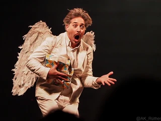 Cirque Bouffon - Célébration  Ein Künstler in weißem Anzug und Engelsflügeln, mit ausdrucksstarker Mimik und offenem Mund.An artist in a white suit and angel wings, with expressive facial expressions and an open mouth.