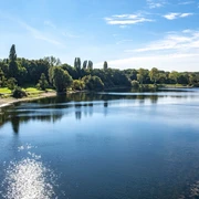 Lake Fühlingen Ein klarer See mit reflektierendem Sonnenlicht und umliegendem Grün unter blauem Himmel in Köln.A clear lake with reflecting sunlight and surrounding greenery under a blue sky in Cologne.