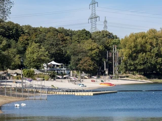 Blackfoot Beach Strand am Fühlinger See vor grünem Wald, mit angrenzendem Café und bunten Booten auf dem Wasser.Beach on Fühlinger See in front of a green forest, with an adjacent café and colorful boats on the water.