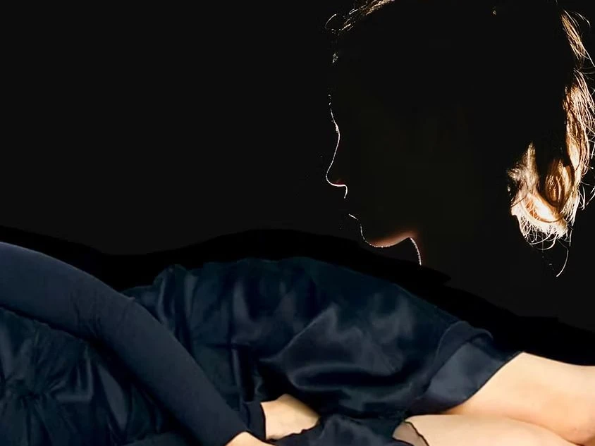 Schatten eines Gesichts und ein liegender Körper in schwarzem Kleid vor tiefschwarzem Hintergrund.