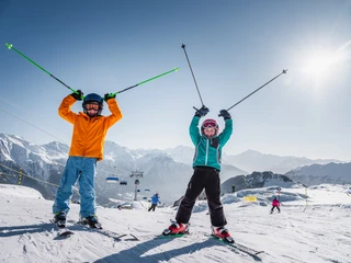 Skispass in der Gruppe unter der Wintersonne Gruppen Skipass Aletsch Arena mit zwei Kindern auf der Skipiste, lachend und mit erhobenen Skistöcken unter strahlender Sonne