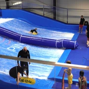 Surfanlage in Pinkys Surfhouse, Mettmann Surfschule mit blauer Indoor-Welle und mehreren Personen, die das Surfen üben.