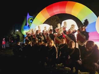 Marschband vor dem beleuchteten Schriftzug Musikgruppe in bunten Uniformen unter leuchtendem Regenbogenbogen bei einer nächtlichen Parade.