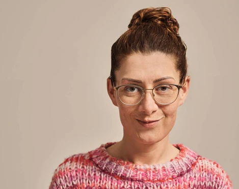 Jana Crämer Ein aufschlussreiches Porträtfoto einer lächelnden Frau mit Brille und buntem Pullover.A revealing portrait photo of a smiling woman with glasses and a colorful sweater.