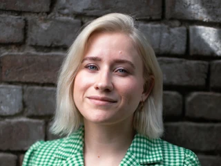 Ruby Braun Eine junge Frau mit kurzem, blondem Haar trägt ein grün-weißes kariertes Jackett und lächelt dezent. Sie steht vor einer gemauerten Steinwand, die im Hintergrund leicht unscharf ist.A young woman with short, blonde hair wears a green and white checked jacket and smiles discreetly. She is standing in front of a brick wall, which is slightly blurred in the background.
