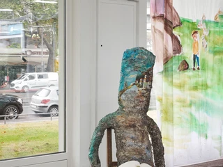 Amelie von Wulffen, installation view Eine surreale Skulptur sitzt auf einem Stuhl in einer Galerie, ein Skizzenbuch auf dem Schoß.A surreal sculpture sits on a chair in a gallery, a sketchbook on its lap.