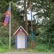 Grenzstation.jpg Kleine bunte Grenzstation mit rot-blauer Schranke und Flagge vor dicht bewachsenem Wald.