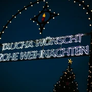 Weihnachtsmarkt Taucha - Weihnachten in der Region Leipzig  Leuchtendes Weihnachtsschild in Taucha bei Nacht, umrahmt von bunten Lichtern und einem Tannenbaum.