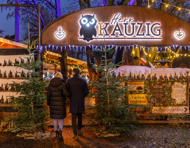 Wintermarkt bei Herr Kauzig - Weihnachten in Leipzig Zwei Personen betreten den Eingang zum kauzigen Wintermarkt im Biergarten von Herrn Kauzig, links und rechts vom Eingang stehen zwei Tannenbäume und im Hintergrund sind festlich beleuchtete Stände zu sehen