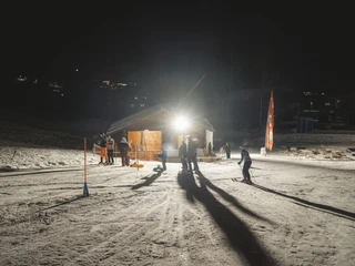 Skilift und Hüttenlicht beim Nachtskifahren im Fieschertal Das Nachtskifahren Fieschertal zeigt den beleuchteten Skilift Blaetz mit Skifahrern und Besuchern im warmen Licht der Aletsch Arena in der Nacht