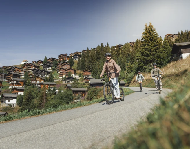 Trottinett-Abfahrt durch das autofreie Dorf Bettmeralp Beim Trottinett Aletsch Arena fahren drei Freunde auf sonnigen Wegen durch das autofreie Bergdorf Bettmeralp mit Blick auf die Walliser Alpen