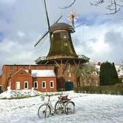 Mühle Pewsum im Winter Zu sehen ist die Mühle Pewsum im Winter, es liegt ein bisschen Schnee, im Vordergrund steht ein altes Fahrrad als Deko.