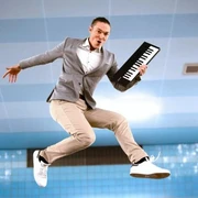 Andreas Langsch Mann in grauem Sakko springt dynamisch in die Luft und hält eine kleine Keyboard-Tastatur.