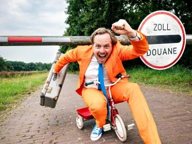 Patrick Nederkoorn (C) Nico Brons Mann im orangefarbenen Anzug auf kleinem Dreirad, lacht, hält Koffer, vor Zollschranke im Freien.