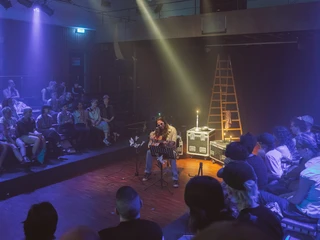 Bonanza festival Eine Person sitzt auf einem Stuhl in einem beleuchteten Raum voller Besucherinnen und Besucher. Im Hintergrund sind Leitern und Beleuchtungstechnik zu sehen.A person sits on a chair in an illuminated room full of visitors. Ladders and lighting equipment can be seen in the background.