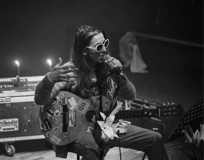 Bonanza festival Eine Person sitzt auf einem Stuhl und spielt Gitarre, während sie singt. Sie trägt eine Sonnenbrille und ist umgeben von Kerzenlicht.A person sits on a chair and plays the guitar while singing. She is wearing sunglasses and is surrounded by candlelight.