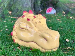 Naturetales premiere: "Alva in the land of shadows" Ein kunstvoll bemalter Drachenkopf ruht auf grünem Gras, hinter einem imposanten Baumstamm.An artistically painted dragon's head rests on green grass behind an imposing tree trunk.