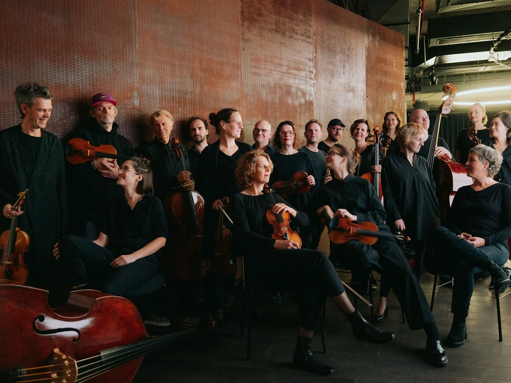 Ensemble Resonanzraum Ein Ensemble von Musiker*innen in schwarzer Kleidung posiert in einem modernen Raum, lächelnd.An ensemble of musicians dressed in black poses in a modern room, smiling.