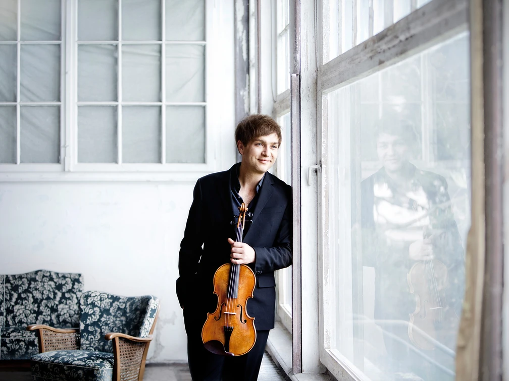 Mönkemeyer, Nils Ein Mann in eleganter Kleidung lehnt lächelnd mit einer Geige in der Hand an einem großen Fenster.A man in elegant clothes leans against a large window with a violin in his hand, smiling.