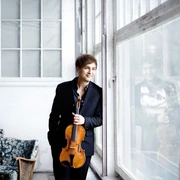 Mönkemeyer, Nils Ein Mann in eleganter Kleidung lehnt lächelnd mit einer Geige in der Hand an einem großen Fenster.A man in elegant clothes leans against a large window with a violin in his hand, smiling.