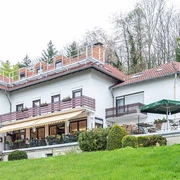 hotel_berghof_galerie_01.jpg Weißes Hotelgebäude mit rotem Dach, großer Terrasse, Sitzplätzen und Blick auf eine begrünte Anhöhe.