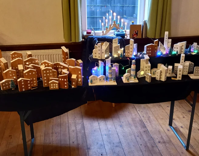 Tisch mit selbstgebastelten Geschenkideen Holzmodelle von Häusern und beleuchtete Stadtsilhouetten auf zwei Tischen in einem hellen Raum.