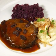 Auf einem Teller ist Sauerbraten mit Rosinen, Kartoffelpürree und Rotkohl angerichtet